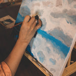 An artist paint a white and blue landscape painting on a canvas and easel.